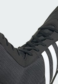 Black athletic shoe with mesh upper, textured finish, white three stripes, and black laces, focusing on a close-up view of the toe area.