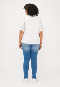 A woman with curly hair faces away, wearing a white top with lace sleeves, blue jeans, and white sneakers against a plain backdrop.