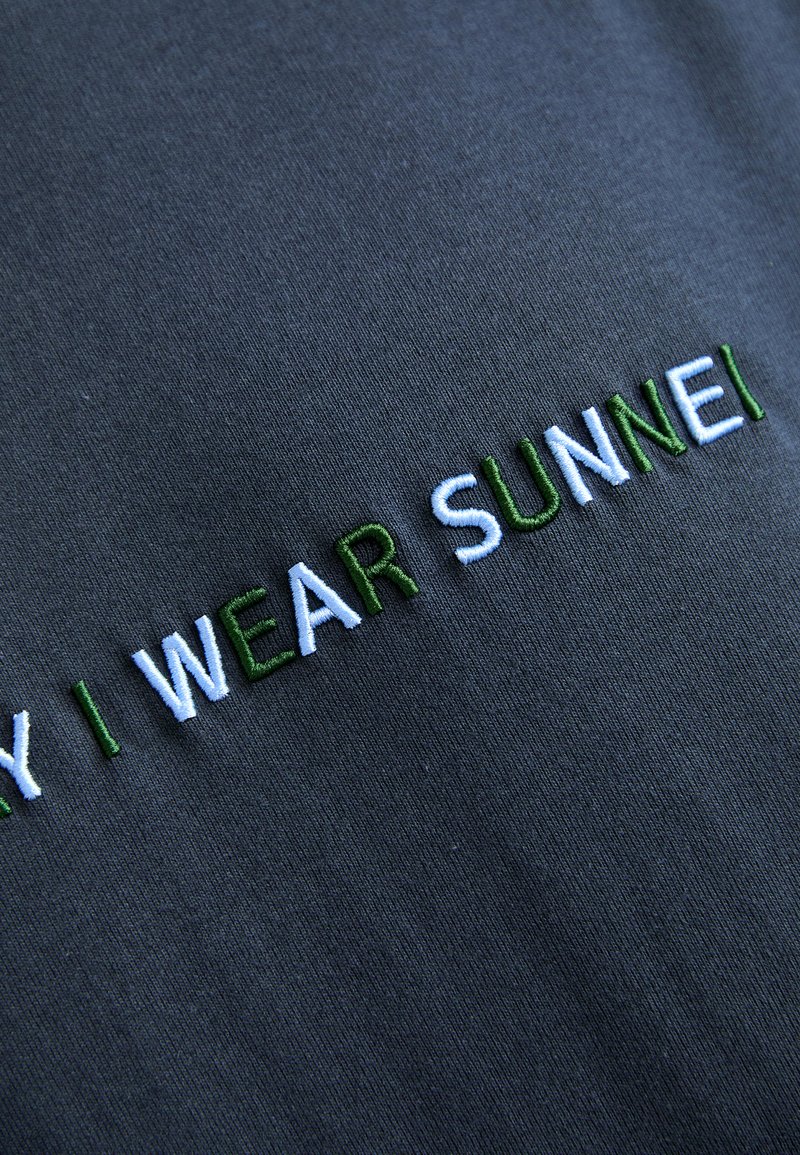 Gray cotton fabric featuring embroidered text in green and light blue reading "WHY I WEAR SUNNEI." Smooth texture with slight sheen.