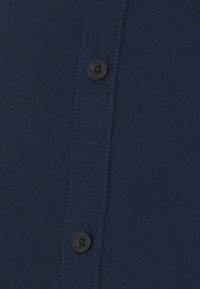 Tessuto di camicia blu scuro con una texture liscia, caratterizzato da bottoni neri lungo la patta centrale e dettagli di cucitura visibili.