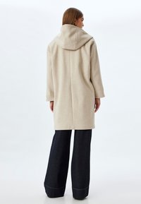 Abrigo de lana beige con capucha, diseño recto, de longitud media y mangas largas. Combinado con pantalones de mezclilla oscuros de pierna ancha.