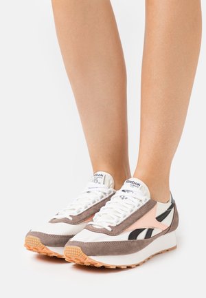 Jambes portant des baskets Reebok Classic blanches, beiges et noires avec semelles en gomme sur un fond blanc.