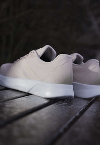 Baskets athlétiques légères beiges avec une tige texturée, trois bandes horizontales et une semelle en caoutchouc blanche. Convient pour une tenue décontractée ou active.