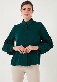 Teal blouse with a button-up front, pointed collar, and puffed long sleeves. Made of smooth fabric, featuring cuff buttons and a relaxed fit.
