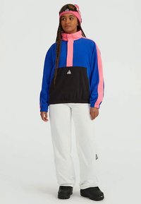 Woman wearing blue, pink, and black ski jacket, white ski pants, black boots, pink beanie, and ski goggles against plain background.