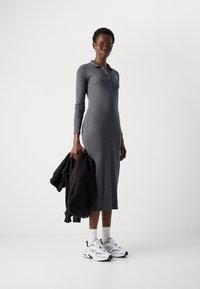 Šedé rebrované maxi šaty s dlhými rukávmi a polo golierom, obsahujúce gombíkovú légou. Čierna mikina držaná v jednej ruke; biele tenisky na nohe.