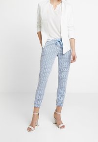 Modré kalhoty s vzorem a šňůrkou v pase, kombinované s bílým blejzrem a topem, a bílými sandály na podpatku. Hladký povrch tkaniny.