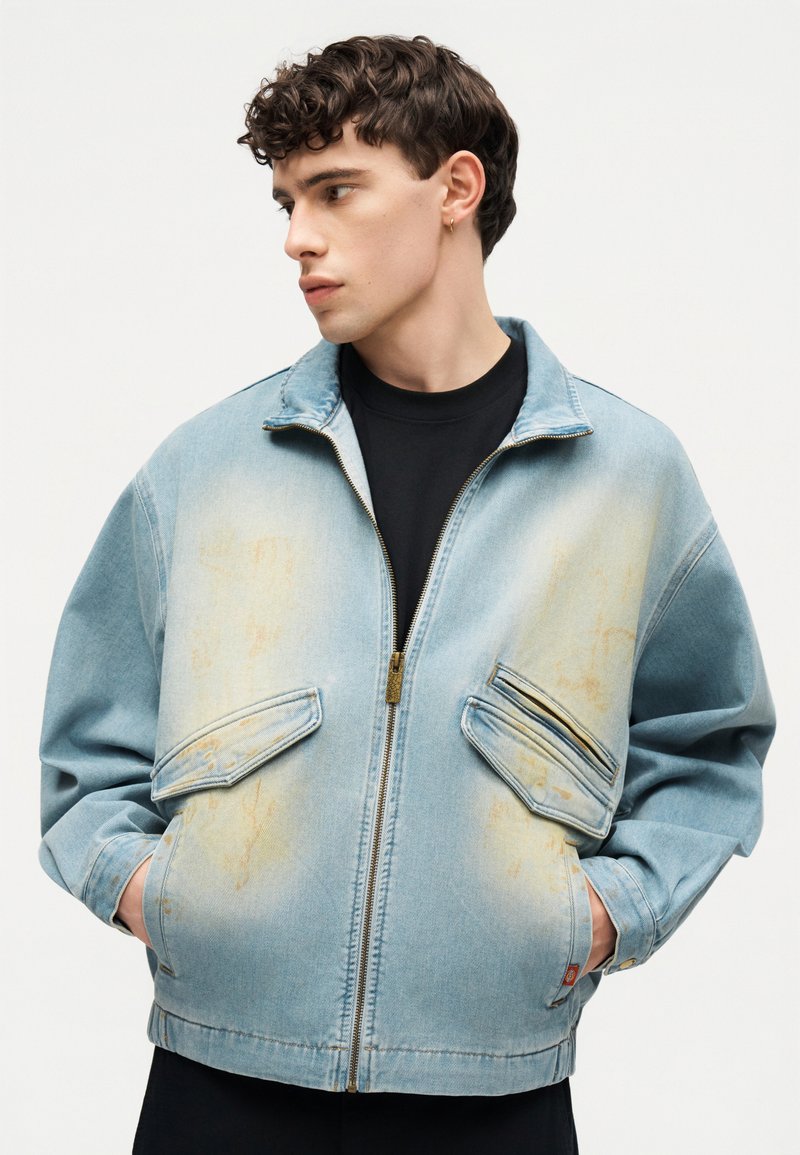 Giovane uomo che guarda a sinistra, mani in tasca, indossa una giacca di denim chiara sbiadita con cerniera e grandi tasche frontali sopra una camicia nera.