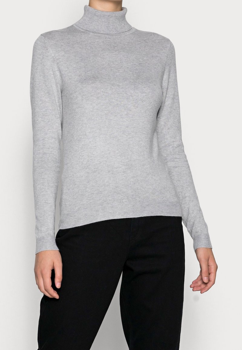 Personne portant un pull à col roulé à manches longues gris clair et un pantalon noir, debout devant un fond clair uni.