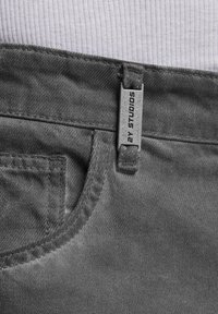 Harmaat farkut, joissa on teksturoidu pinta. Edessä tasku ja vyötärölle kiinnitetty etiketti, jossa on hopeinen brändimerkintä.