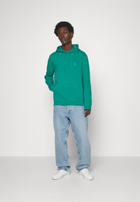 Green cotton hoodie with a front pocket, adjustable drawstrings, and a small logo patch. Worn with light blue jeans and white sneakers.