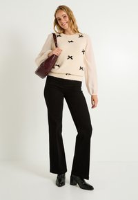 Pull crème avec des accents de nœud noir, manches longues en dentelle ; pantalons noirs évasés ; sac bordeaux ; bottes noires à talons ; sur un fond blanc.