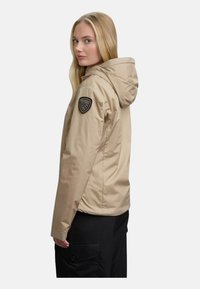 Beige gewatteerde jas met een gestructureerde afwerking, voorzien van een zwart logo-opdruk op de linker schouder en een slim fit silhouet.