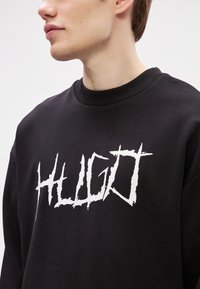 Μαύρο φούτερ με στρογγυλή λαιμόκοψη, με έντονο λευκό σχέδιο λογότυπου. Το ύφασμα φαίνεται απαλό και η εφαρμογή είναι χαλαρή.