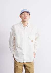 Camicia bianca a maniche lunghe con due tasche frontali, design a bottoni e una toppa con logo. Abbinata a pantaloni beige e un cappellino blu chiaro.