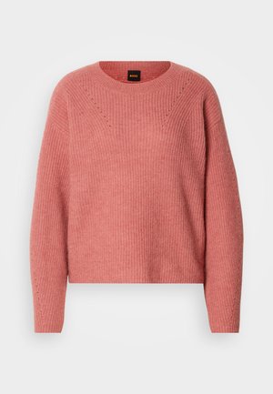 Rozā adīta džemperis ar apaļu kakla izgriezumu, garām piedurknēm un ribotu tekstūru. Iezīmējas ar ševrona rakstu krūtīs un smalkām detaļām.