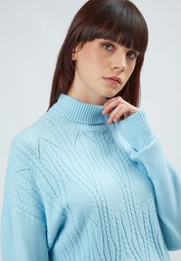 Maglione lavorato a maglia di un azzurro chiaro con un alto colletto a coste. Presenta intricati motivi testurizzati e maniche ampie, evidenziando dettagli raffinati.