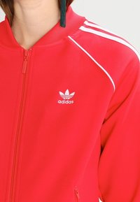 Červená bunda na zip z hladkého polyesteru, s bílými třemi pruhy a malým bílým logem Adidas na hrudi.