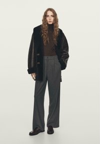 Zwarte nepbonten jas met brede kraag, bruine coltrui, grijze streepjesslip, en donkere schoenen. Model staat tegen een neutrale achtergrond.