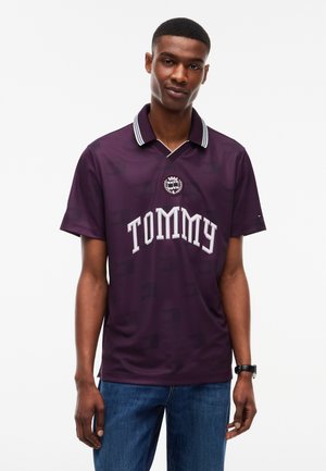 Jeune homme portant un polo violet foncé avec le texte et l'emblème "TOMMY", un jean bleu et une montre noire sur fond blanc.