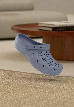 Sabot bleu clair avec des trous de ventilation en forme de fleurs flottant au-dessus d'un tapis beige, près d'une table basse en bois avec des livres empilés.