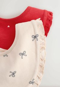 Två ärmlösa toppar: en röd med ribbad halsringning, den andra krämfärgad med broderade rosetter och en volangkant längs ärmhålen.