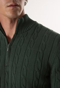 Grüner Kabelstrickpullover mit hohem geripptem Kragen und halbem Reißverschluss. Verfügt über strukturierte Nähte, die das dreidimensionale Design betonen.