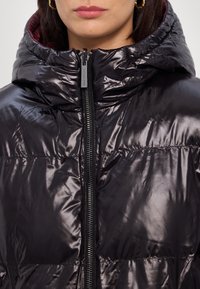 Veste matelassée noire à finition brillante, dotée d'un col montant, d'une fermeture éclair frontale et d'une texture matelassée, doublée de tissu bordeaux.