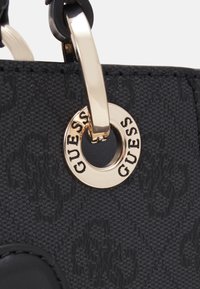 Sac à main noir en tissu texturé, avec des accents brillants et une étiquette en métal ronde gravée "GUESS". Le matériel de fermeture est visible.