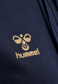 Navyblauwe sweatshirtstof met een gouden Hummel-logo en een bijontwerp, vergezeld van gestileerde lijntjes eronder, met een soepele textuur.