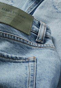 Ljust blå denimtyg har en läderlapp med pr präglad text. Exponerade sömmar och stygn i en kontrasterande nyans ger detaljer.