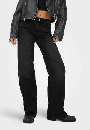 Personne portant un jean noir à jambes larges, un top court noir, une veste en cuir noire et des chaussures noires sur un fond clair uni.