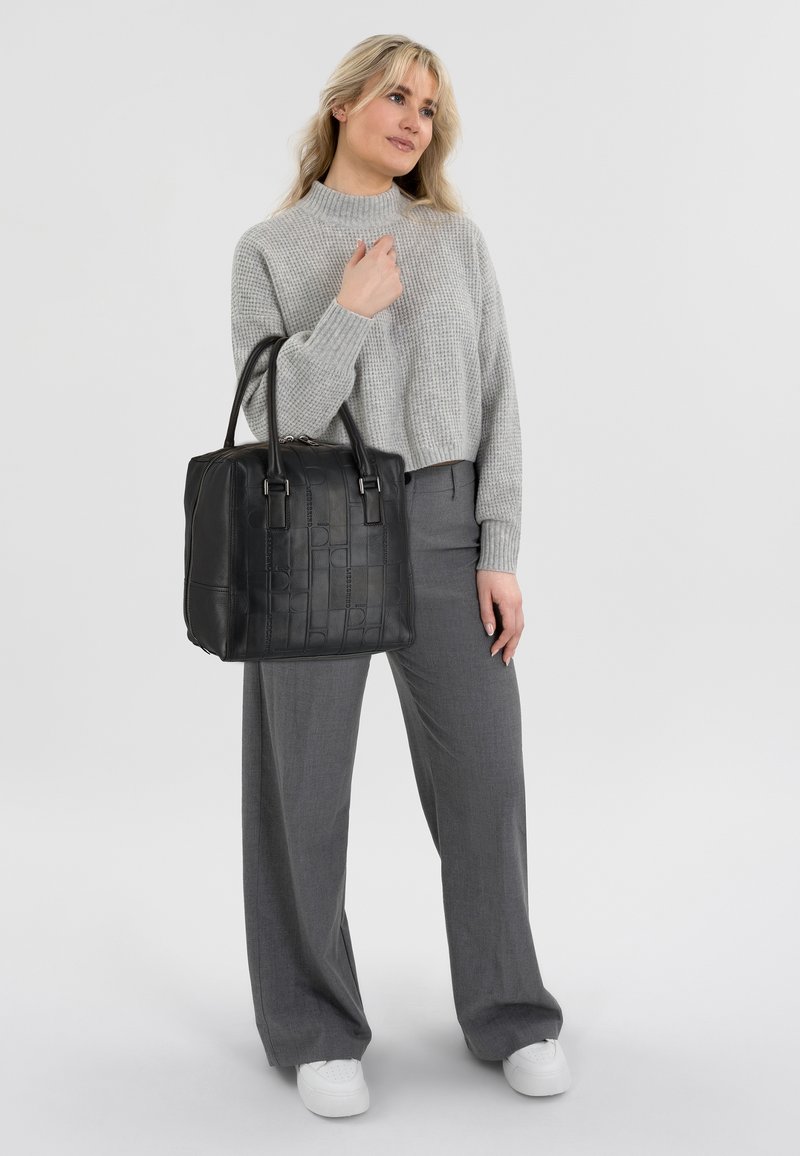 Sac fourre-tout en cuir noir avec un design texturé, doté de poignées doubles et de quincaillerie argentée, aux côtés d'un mannequin portant un pull en maille gris et un pantalon gris.