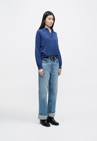 Blauer Strickpullover mit Kragen, kombiniert mit hellblauen Jeans; die Jeans haben aufgerollte Säume und werden durch schwarze Knöchelstiefel ergänzt.