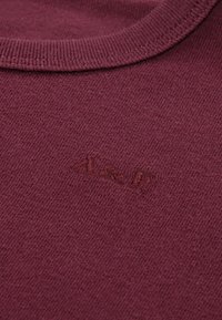 Pull en maille bordeaux avec un col rond. Présente un tissu subtilement texturé et un petit logo brodé près de l'ourlet.
