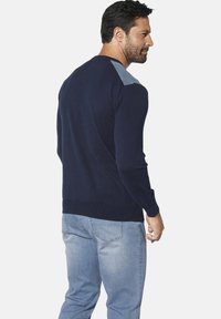 Dunkelblauer Pullover mit langen Ärmeln und geripptem Saum, mit hellgrauen Schulterakzenten und glatter Textur. Tragend mit hellblauen Jeans.