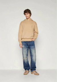 Sweatshirt capuz bege com um bolso frontal, combinado com jeans azuis desbotados com detalhes rasgados, e ténis castanho claro.