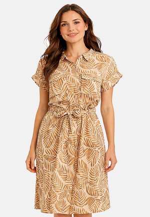Femme portant une robe chemise beige mi-genoux avec manches courtes, poches poitrine, motif feuille et ceinture nouée à la taille, souriante sur fond blanc.