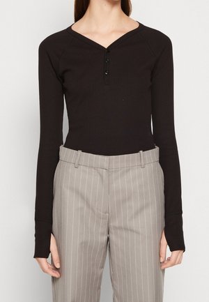 Femme portant un haut henley noir ajusté à manches longues avec trois boutons et un pantalon beige à rayures taille haute.