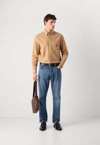 Mann iført en beige skjorte med knapper, blå jeans, svarte sko og svart belte, holder en brun bagg med stropp mot en hvit bakgrunn.