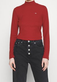 Eine Person trägt einen roten Langarm-Turtleneck und hochtaillierte schwarze Jeans mit vier sichtbaren Knöpfen und steht vor einem schlichten Hintergrund.