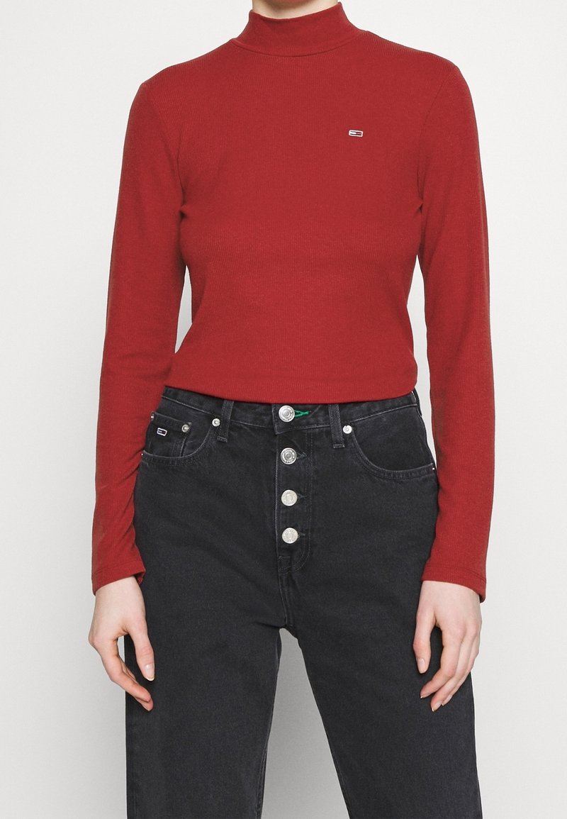 Eine Person trägt einen roten Langarm-Turtleneck und hochtaillierte schwarze Jeans mit vier sichtbaren Knöpfen und steht vor einem schlichten Hintergrund.