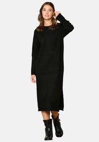 Abito nero a maniche lunghe realizzato in morbido tessuto a maglia, con una vestibilità rilassata, spacco laterale e scollo rotondo. Indossato con stivaletti neri.