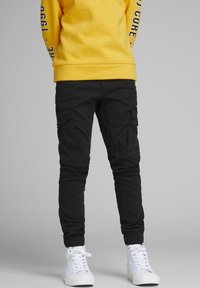 Black cargo pants with multiple side pockets, elastic cuffs, and a smooth texture. Worn with white sneakers and a yellow sweatshirt.