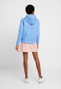 Blå huvtröja med ribbade muddar, ihop med en rosa jeanskjol, bärs med vita sneakers. Bakgrunden är enfärgad vit.