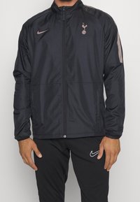 Svart Nike-jacka med hög krage, dragkedja fram och Spurs-logotyp. Har grå detaljer på axlarna och elastiska muddar. Lättviktig material.