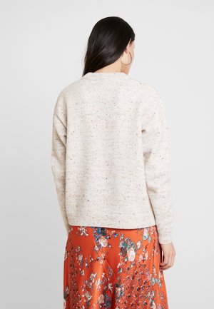 Femme aux longs cheveux noirs portant un pull crème tacheté et une jupe fleurie orange, vue de dos.