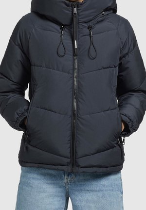 Person iført mørkeblå polstret jakke med frontlynlås og hænder i lommer, kombineret med lyseblå jeans.