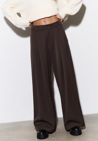 Pantalon marron à jambes larges et taille haute, avec des plis sur le devant et une fermeture à bouton, fabriqué dans un tissu lisse. Chaussures plateformes noires visibles.