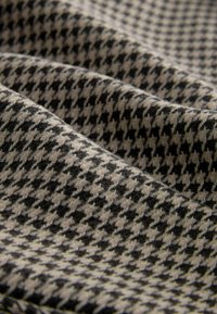 Uldent stof med et sort og beige houndstooth mønster, med synlig tekstur og draperede folder, der skaber dybde.